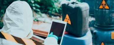 Une personne en combinaison de protection navigue sur une tablette. Un bidon de matière dangereuse est à l'arrière plan